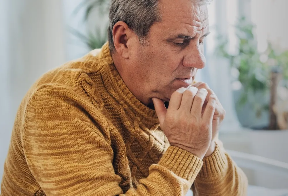 A retired man in a mustard sweater sits with his hands clasped, looking worried as he thinks about his finances.