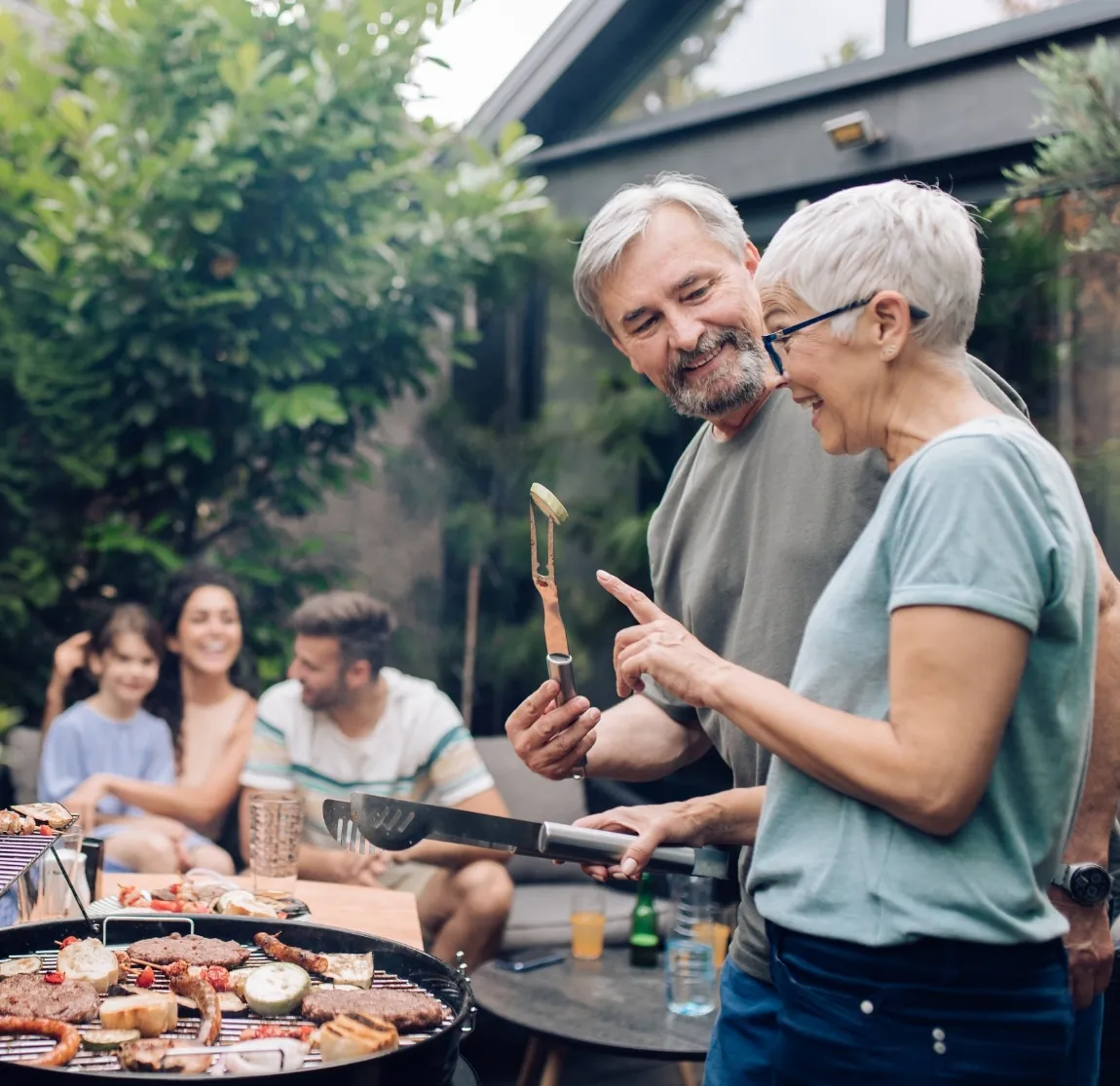 A smiling retired couple grills together at a backyard cookout while family members eat at a table behind them.